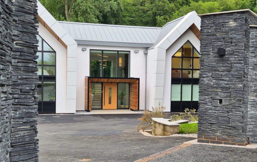 Entrance gates at Woodlands, Draycott in the Clay, Staffordshire. Entrance walls have great slate cladding, the gates are open showing a view of the white house with twin gables and a large central oak wooden door and surround with window above.