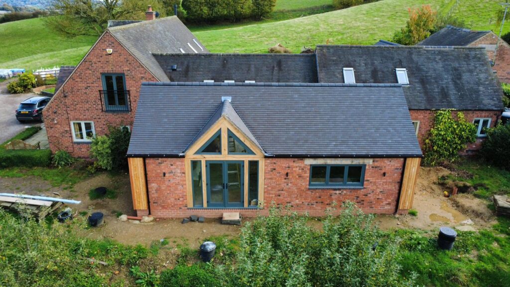 Drone shot of the Herbalshaw Meadow Barn, a house extension in Derbyshire. The image shows a new single-storey brick extension with a dark tiled roof, featuring a striking oak-framed apex entrance and large glass doors, attached to the original red brick barn building. The property is set against rolling green hills.