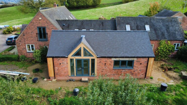 Drone shot of the Herbalshaw Meadow Barn, a house extension in Derbyshire. The image shows a new single-storey brick extension with a dark tiled roof, featuring a striking oak-framed apex entrance and large glass doors, attached to the original red brick barn building. The property is set against rolling green hills.