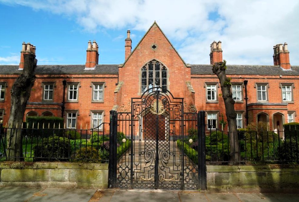 Front view of The Liversage Trust Almshouses on London Road in Derby. Red rick building with large front gable and glass window. Iron gates and railing to the front.