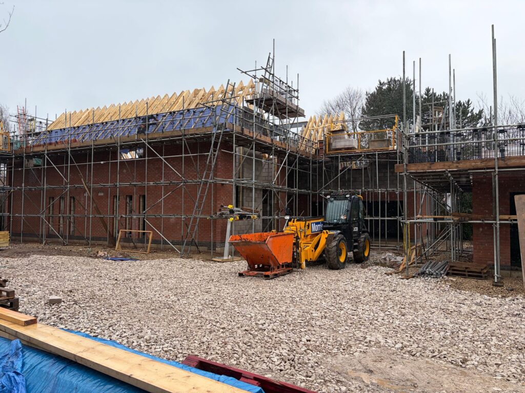 House being constructed. Red brick work and roof trusses being put on. Gravel surrounding the houses and a digger in the foreground.
