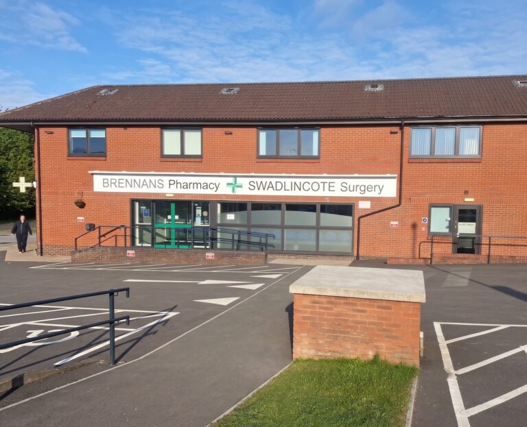 Front view of Swadlincote Doctors Surgery. Double storey brick building, with large glass doors with white sign above.