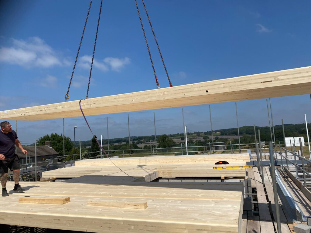 Cross-Laminated Timber First Floor Installed at Locko Road. Cross-Laminated Timber First Floor Installed at Locko Road. Construction worker observing a long engineered wooden beam being lowered by crane chains onto a timber building site surrounded by scaffolding, set against a backdrop of green countryside under a clear sky.